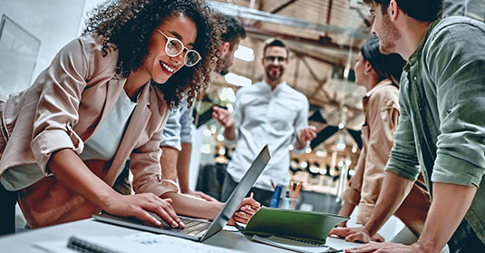 Réunion de bureau, avec en premier plan une femme à lunettes souriante qui utilise son ordinateur