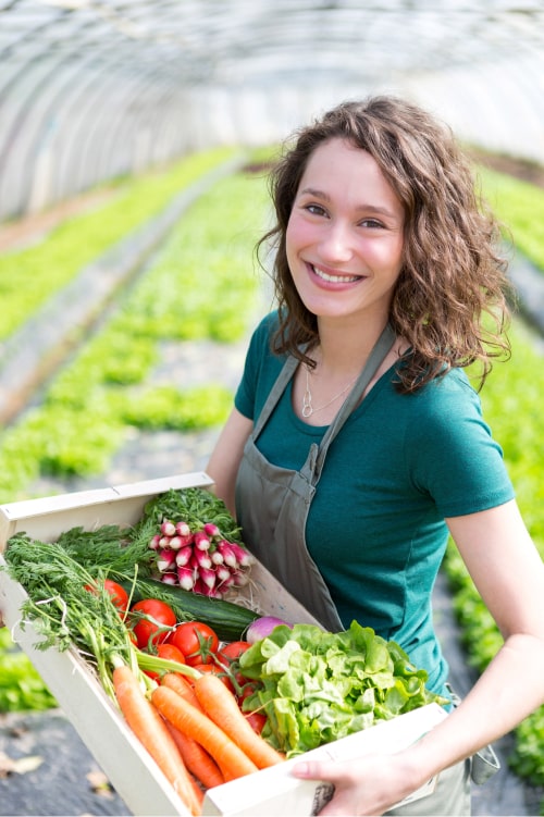 Image d'une agricultrice dans une serre qui tient une cagette de légumes et qui sourit face caméra