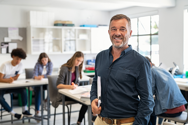 Un instituteur souriant avec une chemise bleu et avec des élèves en fond de salle