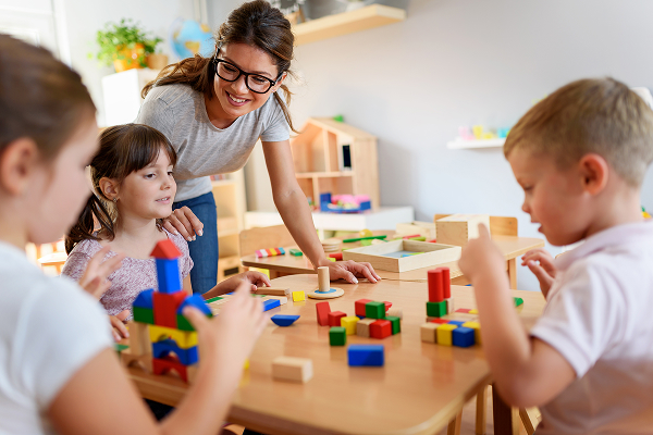 Une jeune institutrice souriante à lunette qui s'occupe de 3 enfants jouant sur une table